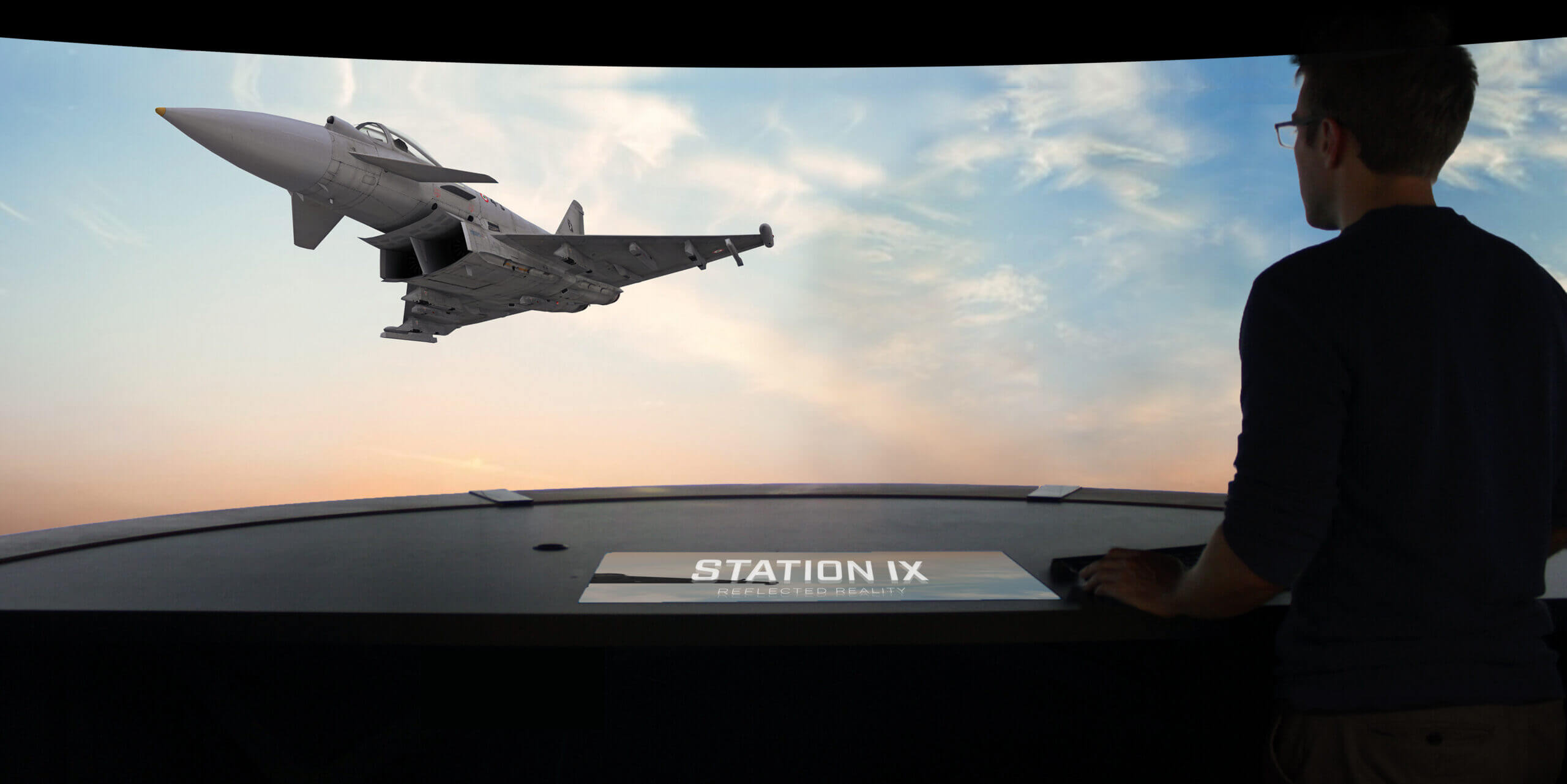 person stands at a control station, observing a fighter jet flying against a colorful sky backdrop.