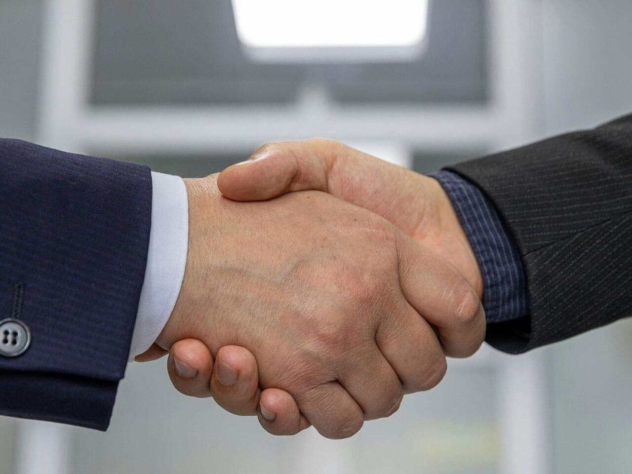 Two hands shake in a business setting, with one wearing a suit and the other in a dress shirt.