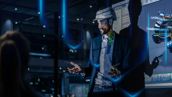 man in a suit and hard hat gestures while presenting data on a screen in a dimly lit office environment.