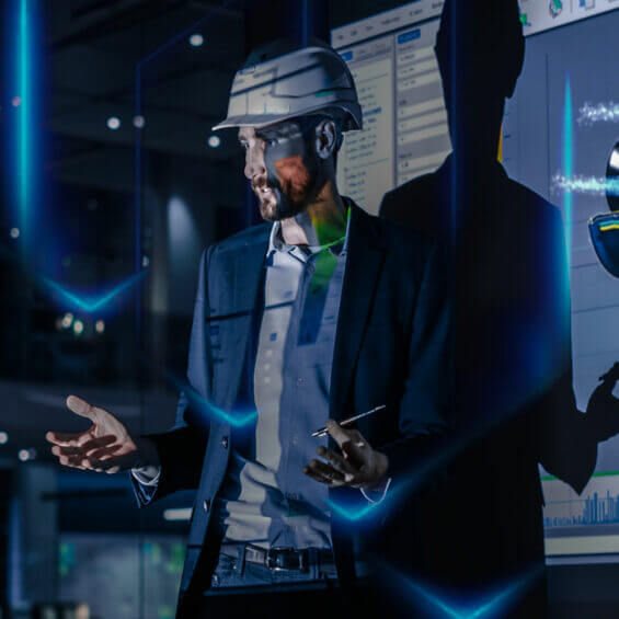 man in a suit and hard hat gestures while speaking in front of a digital screen displaying data and graphics.