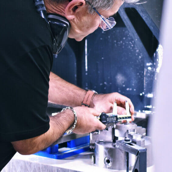 person measures a metal part with calipers in a workshop, wearing safety glasses and ear protection.